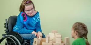 Picture of A young handicap girl is sitting in her wheelchair and is playing with other children - they are building towers using wooden blocks.