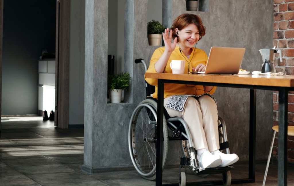 Picture of a woman in a wheelchair using her laptop at a work table smiling and waving.