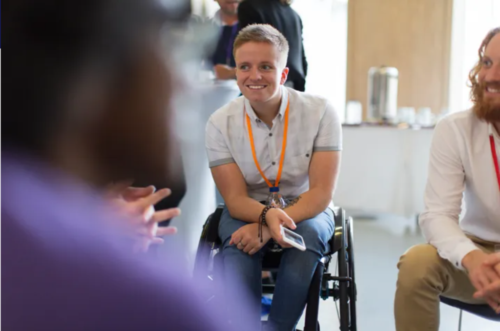Image of a young male sitting in his wheelchair with a group of people holding a phone wearing a bright orange lanyard.