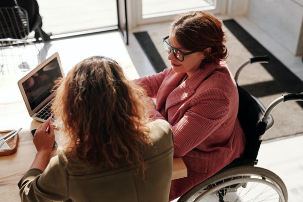 Woman in Wheelchair with woman seated at table looking at a laptop together in a sunny room.