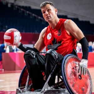 Mike Whitehead in a red jersey sitting in a modified wheelchair holding a ball in his right hand and looking very determined into the camera lens.