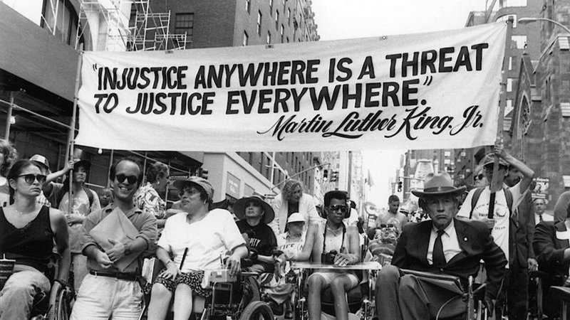 Black and White image of a protest parade with a line of people in wheelchairs advancing with banner reading Injustice Anywhere is a Threat To Justice Everywhere 