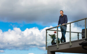 Chuck Edward standing on a balcony with a partly cloudy sky behind him.