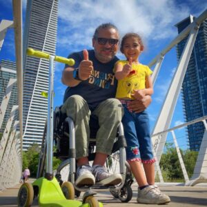 A man in a wheel chair smiling with thumbs up with a young girl standing next to him