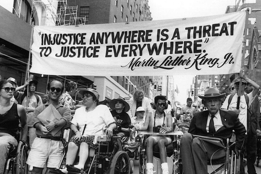 Judy Heumann is sitting in the street with other people using wheelchairs. They are protesting and carrying a banner that reads, "Injustice anywhere is a threat to justice everywhere. Martin Luther King Jr."