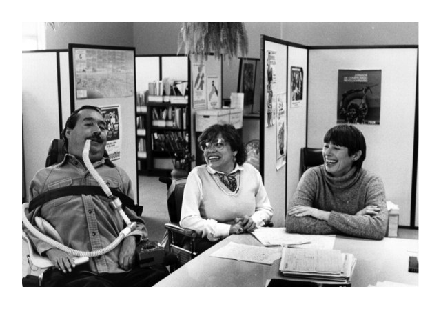 Judy Heumann sits in an office laughing with man using a wheel chair and another woman sitting in a chair.