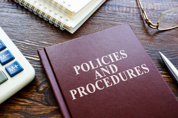 A burgundy book sits on a wooden table. It is titled, Policies and Procedures.