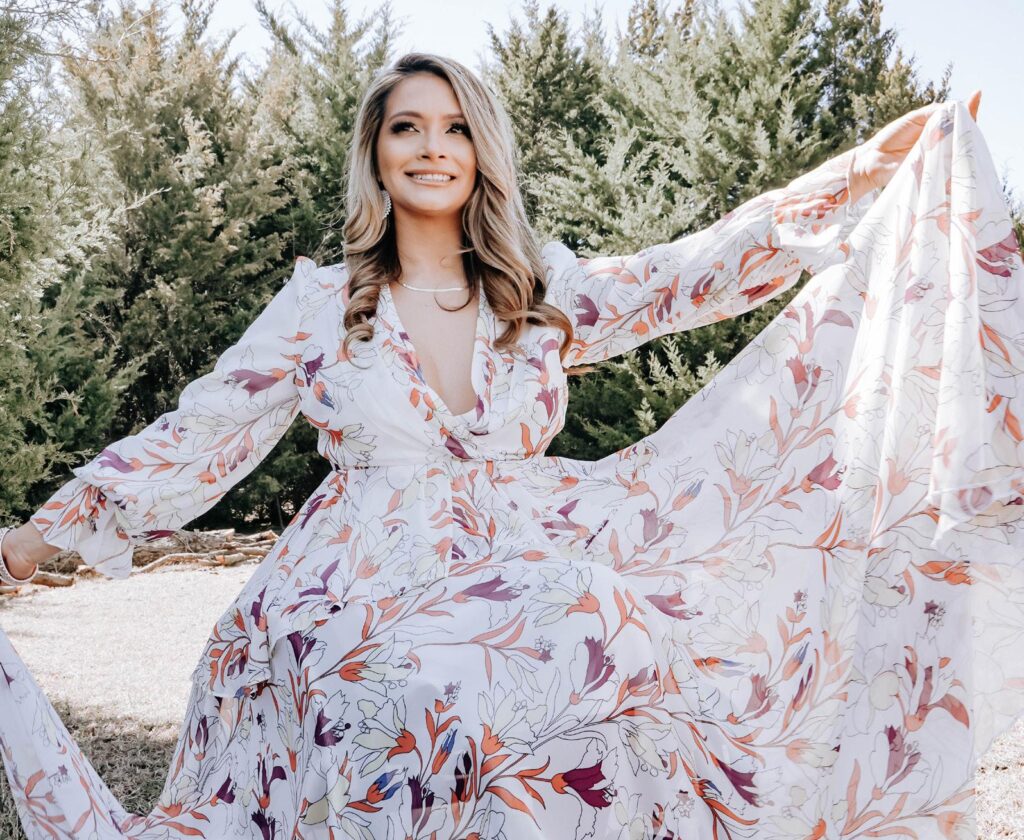 A portrait of Marcela Marañon smiling outdoors in front of pine trees holding up the bottom of her floral white dress.